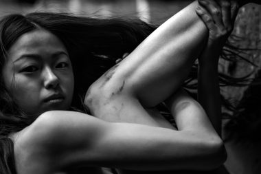 Black and white photo of a young Asian woman hugging her legs and looking at the camera.