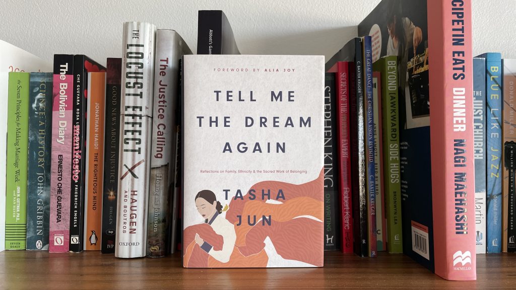 "Tell Me The Dream Again" by Tasha Jun, on a bookshelt in front of the spines of other books.