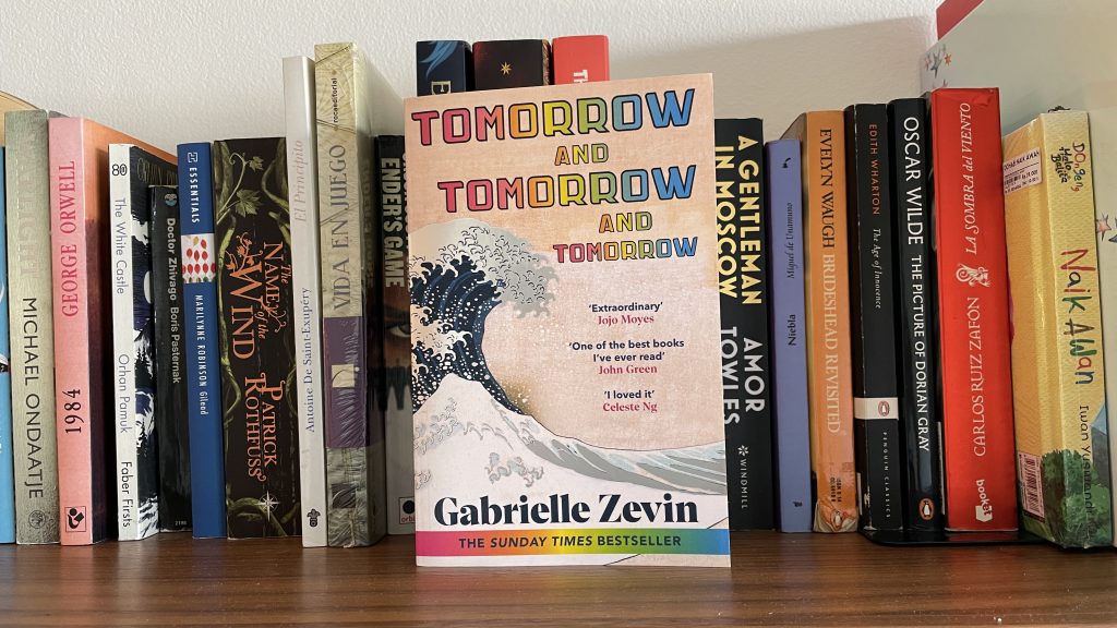 "Tomorrow, and Tomorrow, and Tomorrow" by Gabrielle Zevin, on a bookshelt in front of the spines of other books.