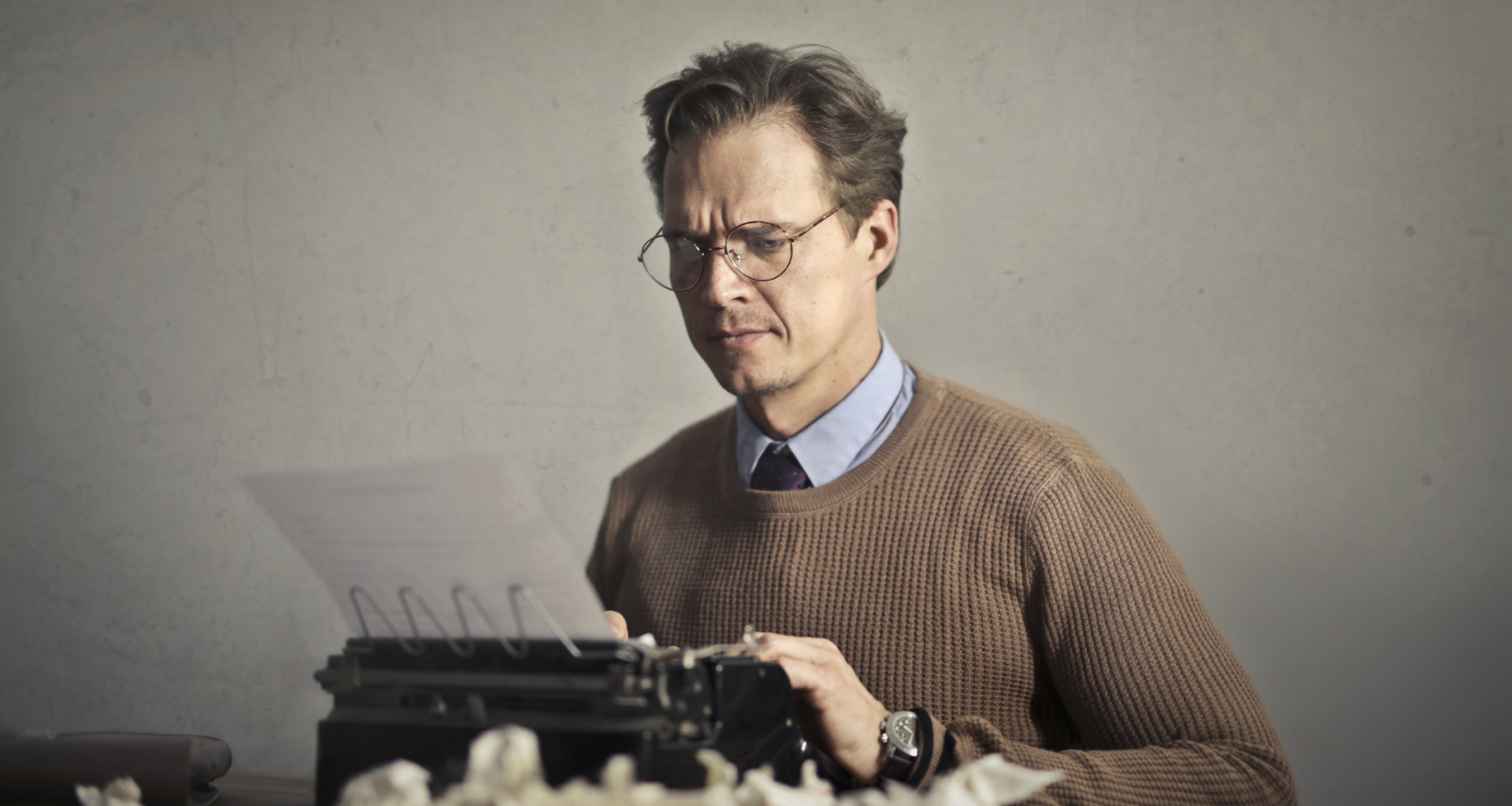 Middle-aged white man in a collared shirt and sweater looks intently at page with his fingers on a typewriter and crumpled paper in front.